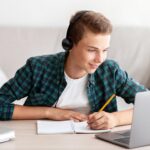 Joyful,Teen,Guy,Writing,At,Notebook,And,Looking,At,Laptop